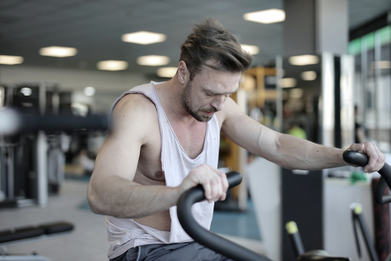 A man wearing a white tank top and gray shorts is using a black fan bike inside the gym