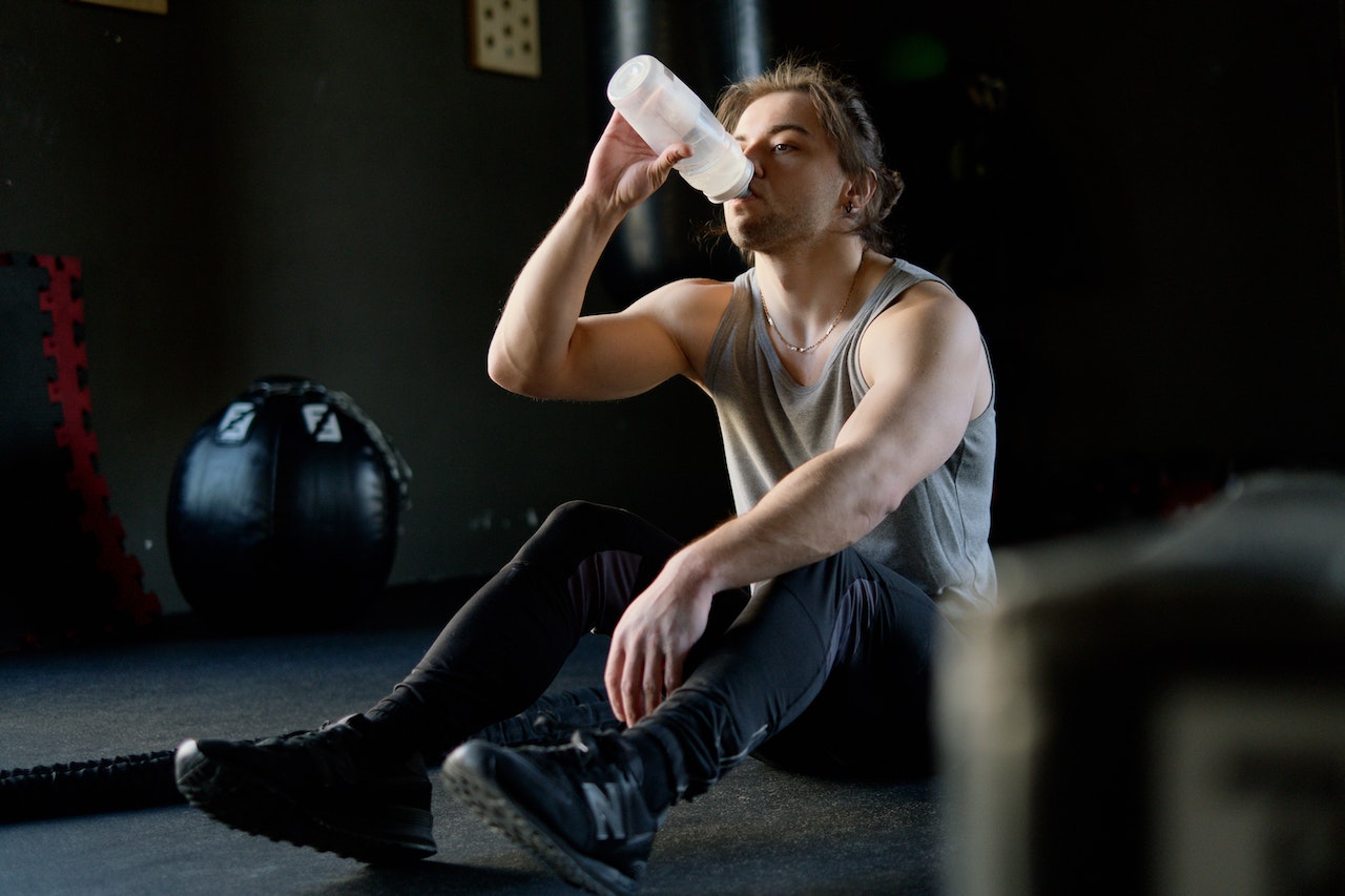 A man wearing a gray tank top and black leggings is drinking from a transparent tumbler