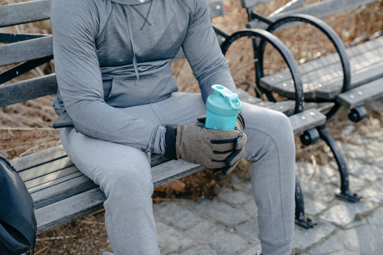 A person wearing a gray hoodie and gray sweatpants is sitting on a wooden bench while holding a blue tumbler