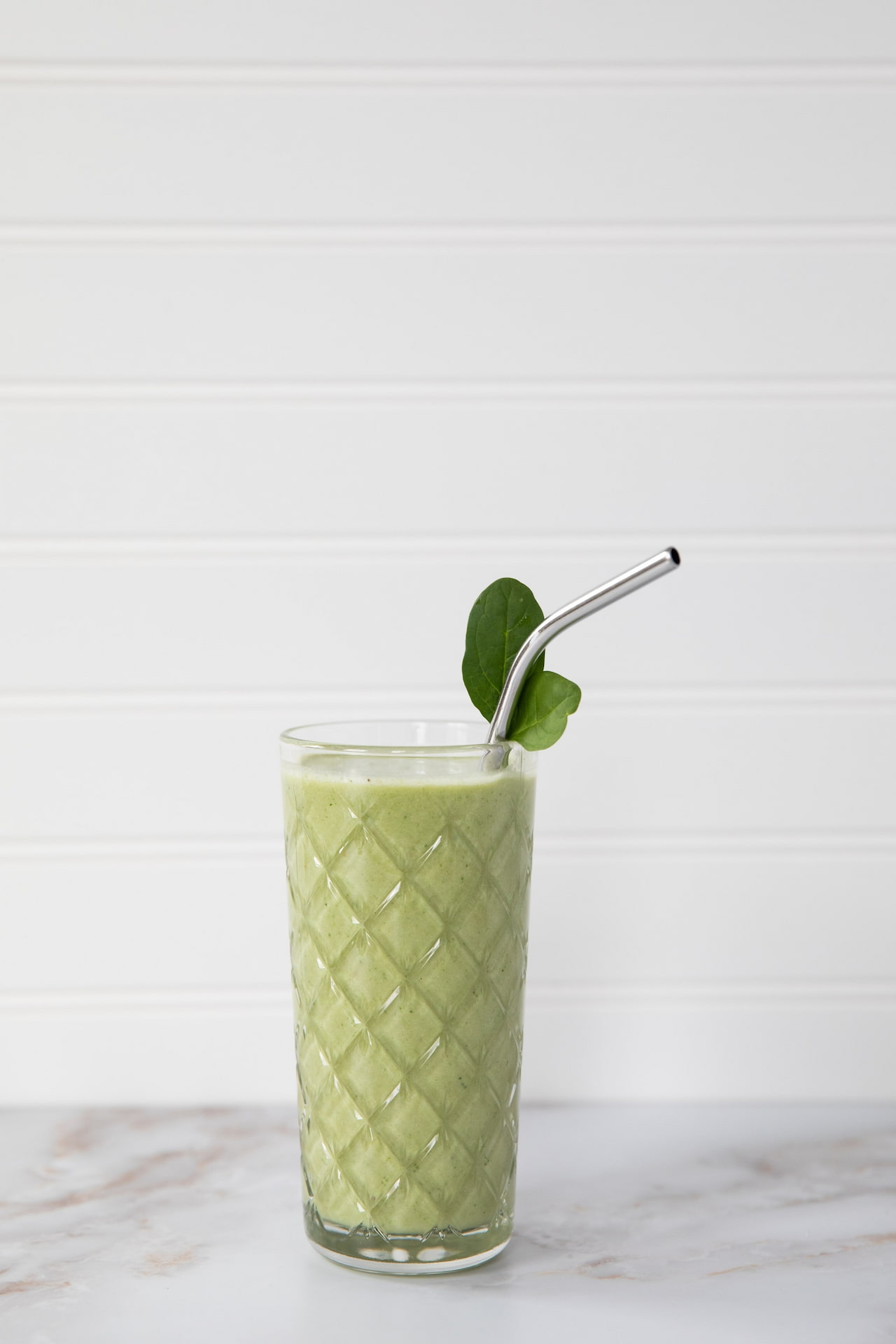 A clear glass of green shake with a metallic straw near a white wall