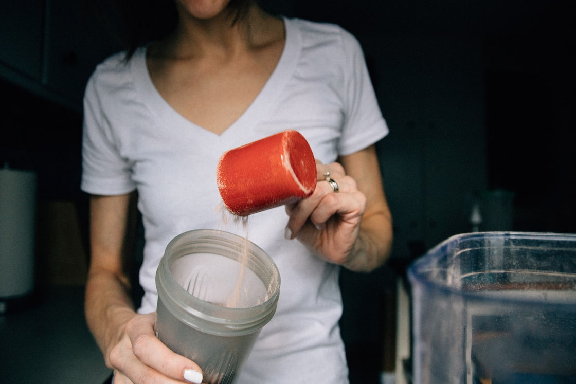 A woman wearing a white v-neck shirt is using a red scoop to put protein powder in a clear plastic tumbler