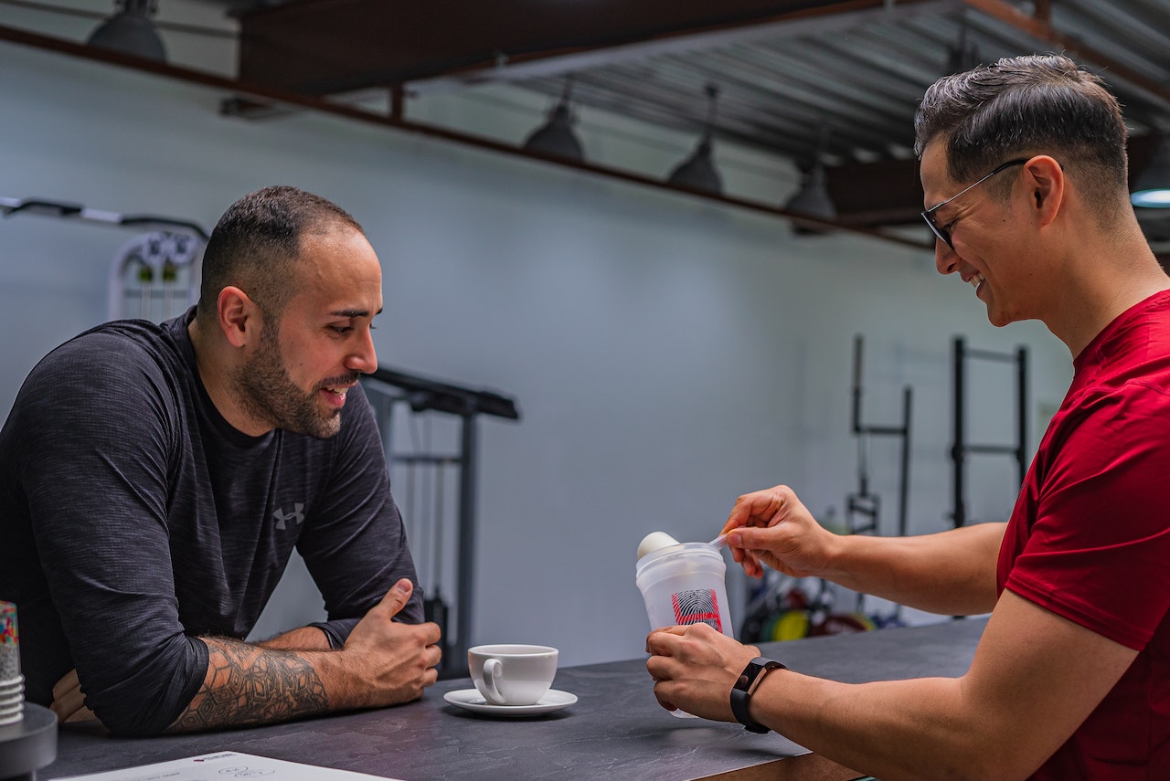 A man wearing a red shirt is using a white scoop to make a protein drink in front of a person wearing a black long-sleeve shirt