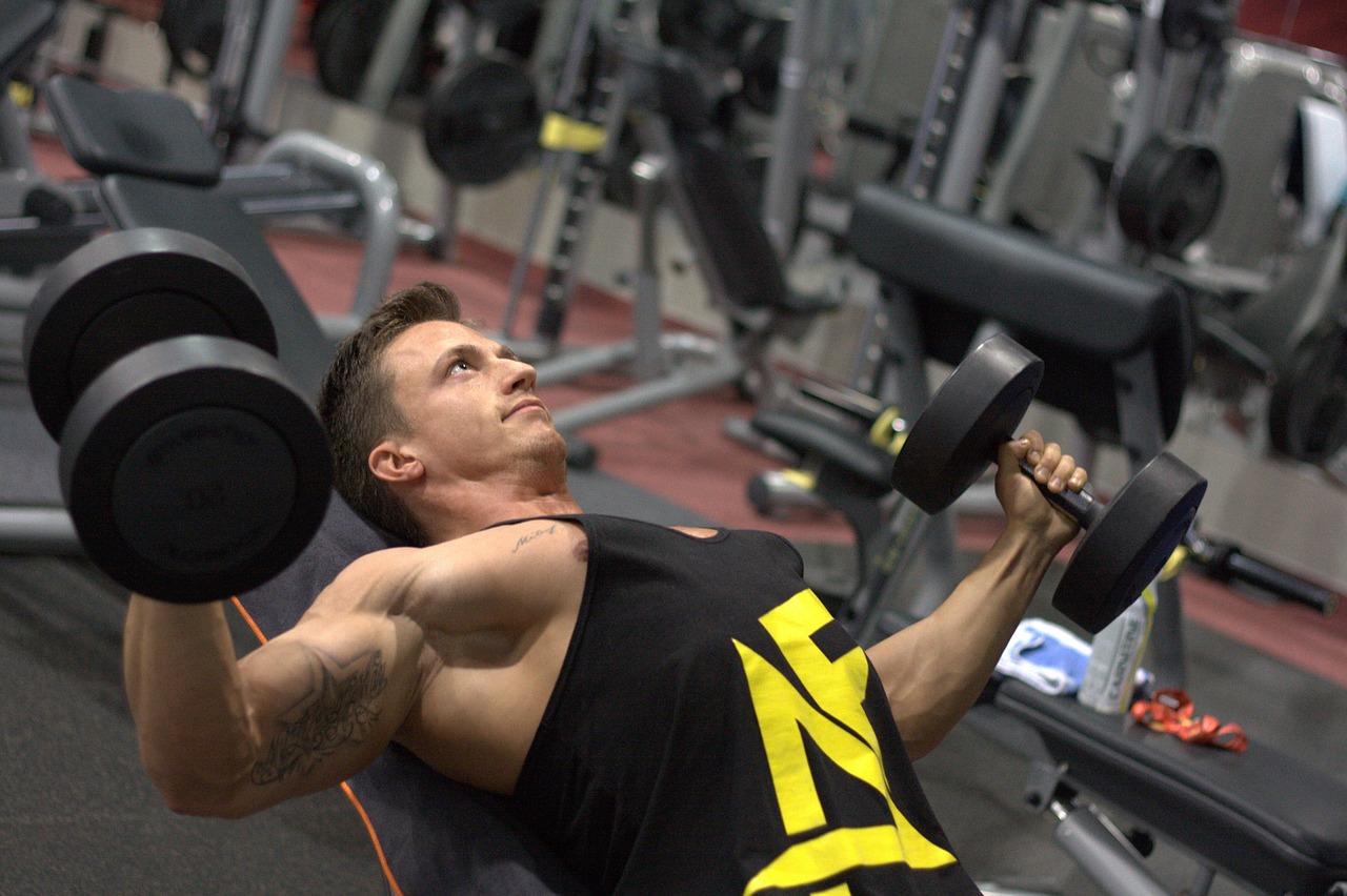 A man wearing a black tank top is lying down on black gym equipment while lifting two black dumbbells