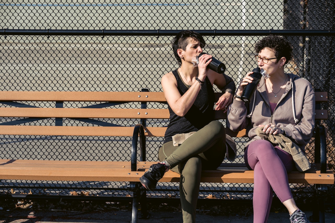 A woman wearing a black tank top and green leggings and a woman wearing a brown hoodie and purple leggings are sitting on a brown wooden bench