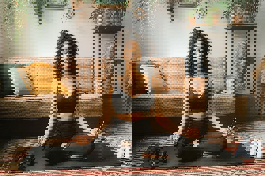Woman doing a yoga pose in her living room while her dumbbells. water bottle and shoes are placed around her