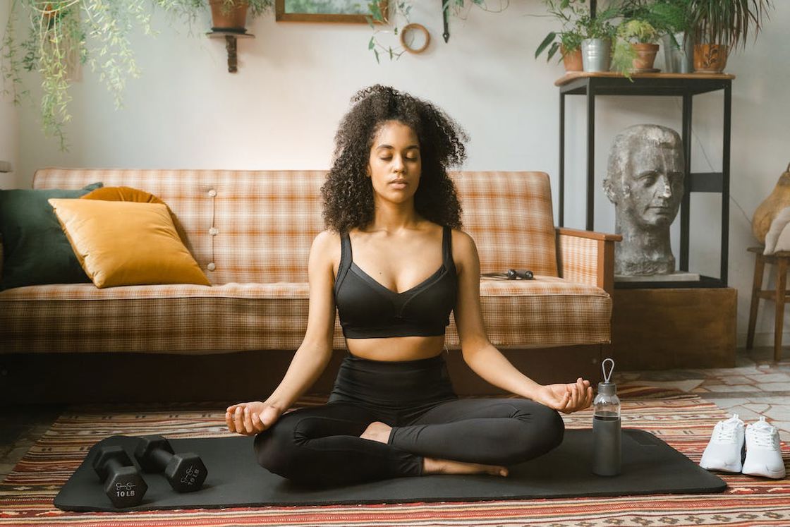 Woman doing a yoga pose in her living room while her dumbbells. water bottle and shoes are placed around her