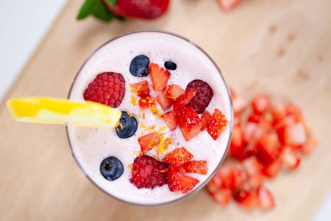 Freshly cut raspberries, blueberries, and strawberries on top of a smoothie on a clear glass placed on a brown wooden table