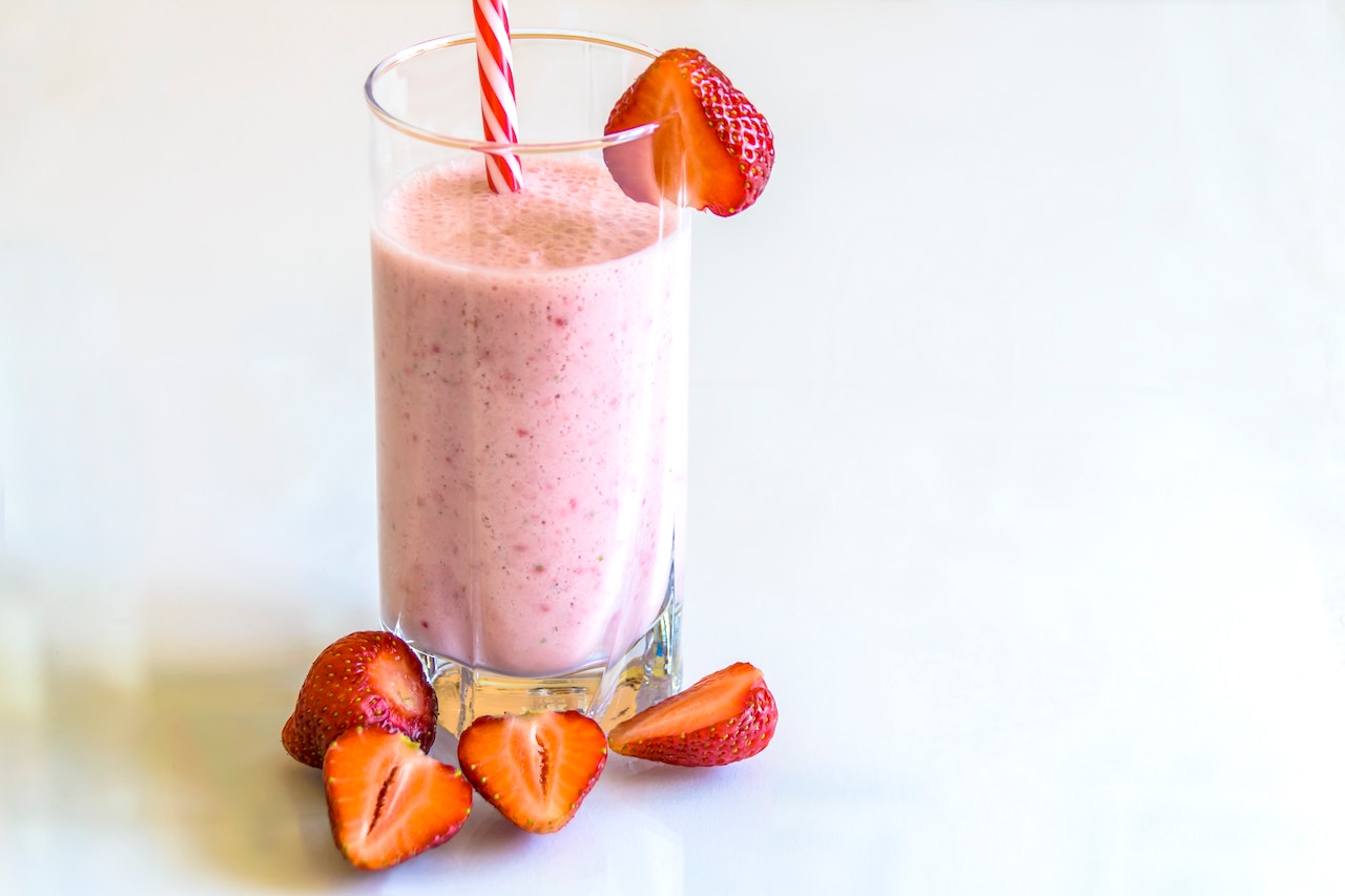 A strawberry protein smoothie on a tall clear glass with plastic straws and freshly cut strawberries on top of a white surface