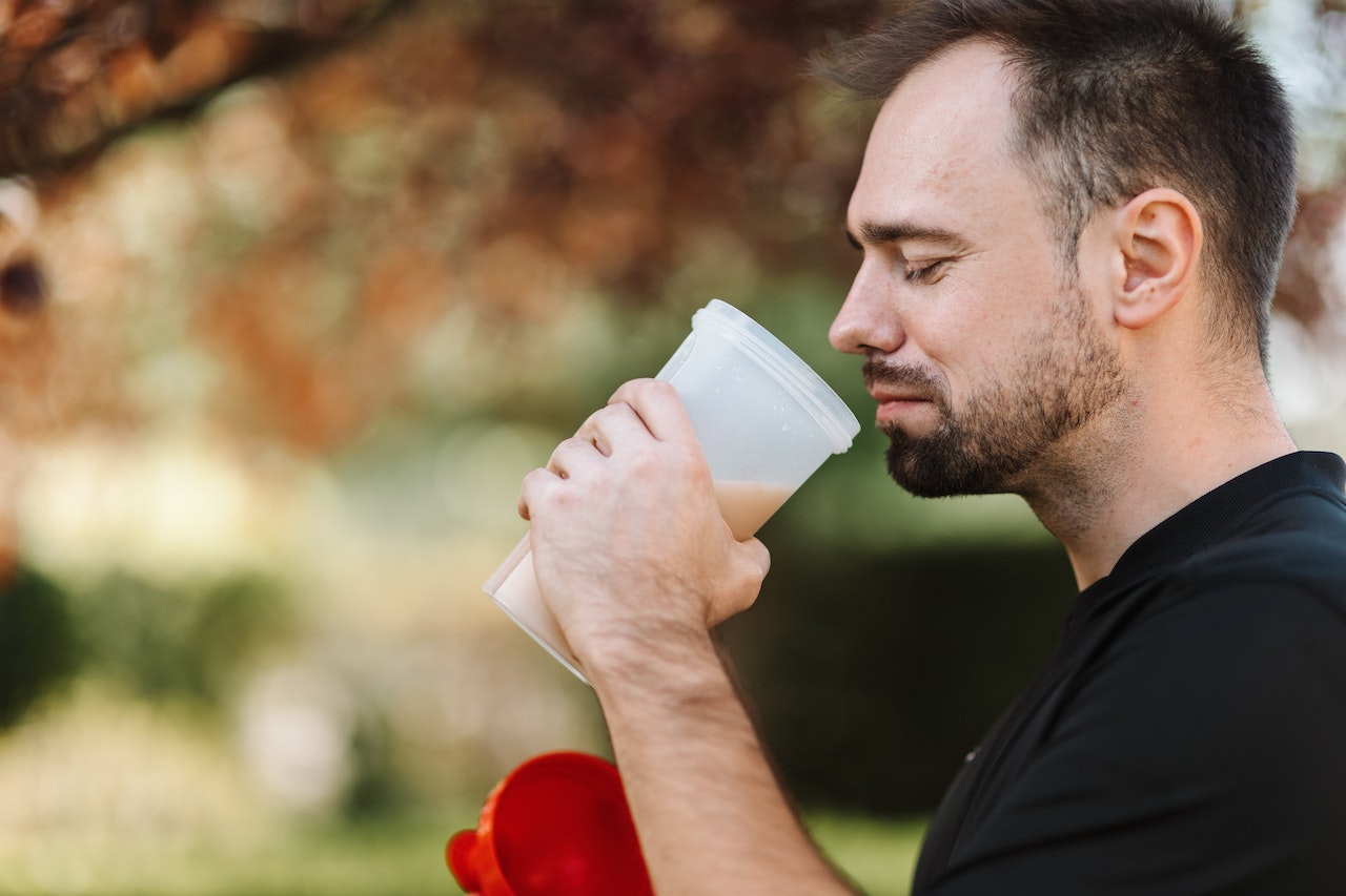 A man wearing a black shirt is holding a plastic tumbler with a red lid filled with a brown protein drink