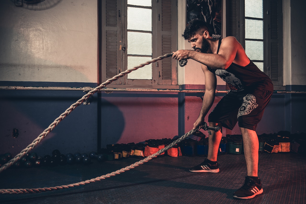 A man wearing a black tank top and black shorts is using the brown battle rope inside the gym