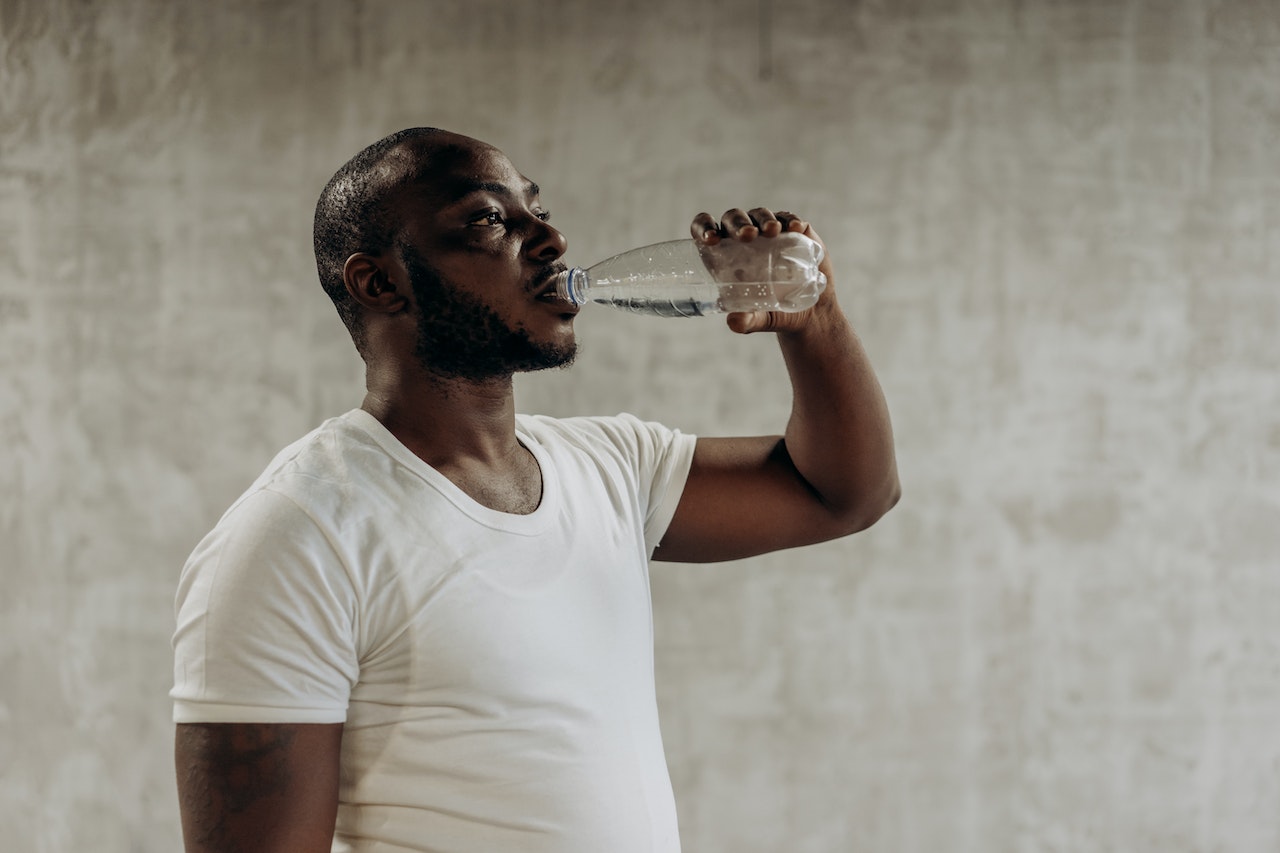 A man wearing a white shirt over a white tank top is drinking water from a plastic bottle near a gray wall